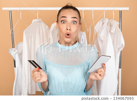 Cashback Concept. Portrait of surprised excited woman holding smartphone and credit card in hands, shocked by seasonal sales and discounts, ordering and buying stylish clothes and purchases online Cashback Concept. Portrait of surprised excited woman holding smartphone and credit card in hands, shocked by seasonal sales and discounts, ordering and buying stylish clothes and purchases online 134067093