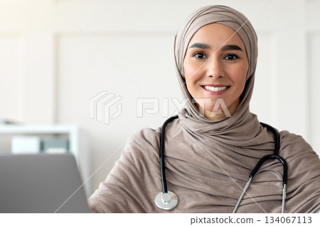 Beautiful muslim woman in hijab doctor with stethoscope posing in hospital, sitting at worktable with laptop, medical folder, smiling at camera, copy space. Doctor's office, cabinet interior concept Beautiful muslim woman in hijab doctor with stethoscope posing in hospital, sitting at worktable with laptop, medical folder, smiling at camera, copy space. Doctor's office, cabinet interior concept 134067113