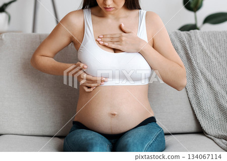Breast lumps in pregnant women. Young expectant mother suffering painful feelings in chest area, worried millennial lady in white top bra massaging herself while sitting on couch at home, free space 134067114