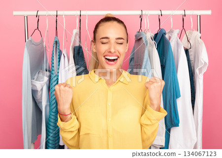 Sales And Discounts. Portrait Of Emotional Young Lady Screaming Yes With Joy, Closing Eyes, Shaking Clenched Fists. Female Customer Laughing And Gesturing, Clothing Rail In The Background 134067123