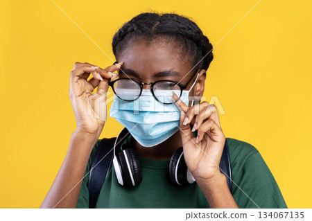 Don't Touch Your Face. Black Female Student Wearing Medical Mask And Scratching Eye Standing Over Yellow Studio Background. Covid-19 Pandemic Outbreak, Coronavirus Protection 134067135