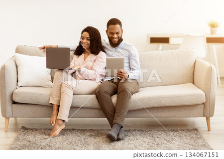 Weekend Leisure. Portrait Of Happy Millennial Black Couple Resting On Sofa. Man Using Digital Tablet, Woman Working On Laptop Computer. Family Browsing Internet Together Sitting On Couch 134067151