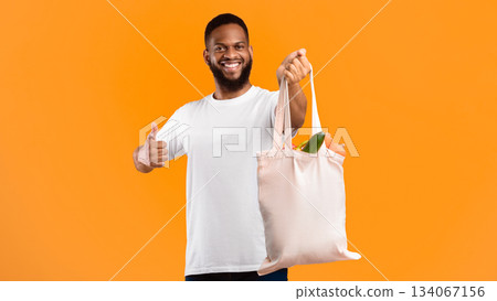Plastic-Free Life. African Man Holding Eco Bag With Groceries And Gesturing Like Smiling To Camera Standing Over Yellow Studio Background. I Like Eco-Friendly Lifestyle, Ecology And Consumerism 134067156