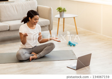 Smiling African American Woman Having Online Fitness Training With Laptop At Home, Using Computer And Talking With Coach, Exercising On Yoga Mat In Living Room, Enjoying Healthy Lifestyle 134067159