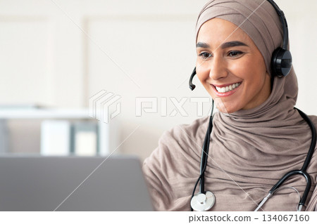 Cheerful muslim lady doctor having video chat with patient, using headset and laptop, talking to sick client online while working at clinic, copy space. Female physician having online appointment 134067160