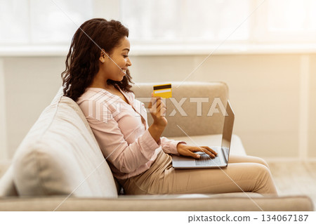 Easy Payment And Cashback Concept. Side view portrait of smiling african american young woman holding credit card in hand and using laptop, sitting on couch at home, typing on keyboard, copy space Easy Payment And Cashback Concept. Side view portrait of smiling african american young woman holding credit card in hand and using laptop, sitting on couch at home, typing on keyboard, copy space 134067187