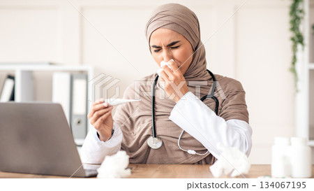 Ill doctor young muslim woman in hijab sneezing nose and checking fever while sitting at her workdesk in front of laptop, got sick during flu or COVID-19 pandemic, clinic interior Ill doctor young muslim woman in hijab sneezing nose and checking fever while sitting at her workdesk in front of laptop, got sick during flu or COVID-19 pandemic, clinic interior 134067195