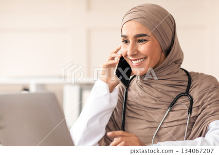 Cheerful muslim lady doctor having conversation on mobile phone with patient, looking at laptop screen, talking to sick client while working at clinic, copy space. Female physician having appointment Cheerful muslim lady doctor having conversation on mobile phone with patient, looking at laptop screen, talking to sick client while working at clinic, copy space. Female physician having appointment 134067207