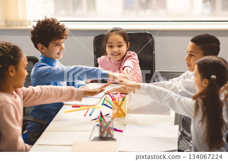 Youth Collaboration Concept. Group of multiethnic elementary schoolchildren sitting in circle at table and putting hands together in stack, having fun. Friends and fellowship, partnership and support Youth Collaboration Concept. Group of multiethnic elementary schoolchildren sitting in circle at table and putting hands together in stack, having fun. Friends and fellowship, partnership and support 134067224