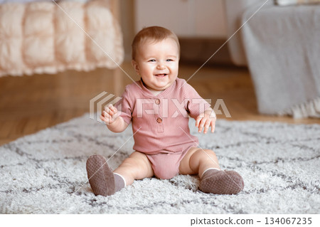 Happy little baby boy or girl sitting on carpet at home, smiling at camera, living room interior, copy space. Adorable toddler having fun, sitting on floor and laughing, discovering world Happy little baby boy or girl sitting on carpet at home, smiling at camera, living room interior, copy space. Adorable toddler having fun, sitting on floor and laughing, discovering world 134067235