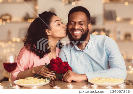 Celebrating Special Day. Beautiful black girlfriend kissing her smiling boyfriend in cheek, holding bouquet of red roses. Couple sitting at table in luxury restaurant during romantic date and dinner Celebrating Special Day. Beautiful black girlfriend kissing her smiling boyfriend in cheek, holding bouquet of red roses. Couple sitting at table in luxury restaurant during romantic date and dinner 134067236
