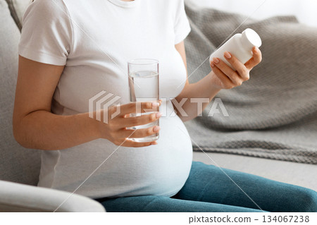 Prenatal Vitamins. Portrait Of Beautiful Smiling Pregnant Woman Holding White Jar With Pills And Glass Of Water, Taking Supplements For Healthy Pregnancy While Sitting On Couch At Home, Free Space 134067238