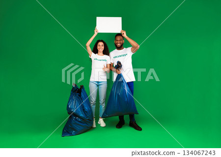Two Diverse Volunteers Holding Blank Paper Board Posing With Plastic Bags Full Of Wasted Junk Standing Over Green Studio Background. Volunteering Work Concept. Mockup Two Diverse Volunteers Holding Blank Paper Board Posing With Plastic Bags Full Of Wasted Junk Standing Over Green Studio Background. Volunteering Work Concept. Mockup 134067243