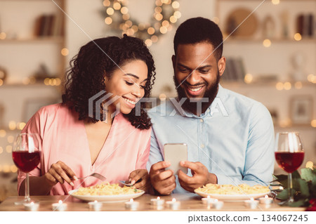 Happy Loving Family. Beautiful young african american couple using cellphone, sitting at table and having dinner, eating pasta. Smiling man sharing photos with his woman, spending time together 134067245
