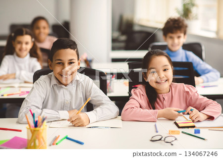 Education And Children Concept. Portrait of happy smiling diligent boy and girl sitting at table in classroom at elementary school with group of classmates in background and posing, looking at camera 134067246