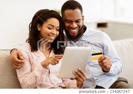 Online Shopping And Ecommerce. Happy young african american family of two people sitting on sofa at home, choosing and buying clothes or products online, paying with credit card using digital tablet 134067247
