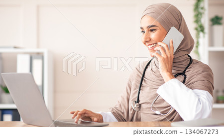 Beautiful muslim woman in hijab doctor looking at laptop screen while talking on mobile phone, having conversation with sick patient, giving recommendations, prescribing treatment on phone, free space Beautiful muslim woman in hijab doctor looking at laptop screen while talking on mobile phone, having conversation with sick patient, giving recommendations, prescribing treatment on phone, free space 134067273