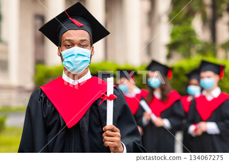 Black guy student in graduation costume showing diploma at camera, posing over international group of students at university campus, wearing protective face mask, panorama with copy space Black guy student in graduation costume showing diploma at camera, posing over international group of students at university campus, wearing protective face mask, panorama with copy space 134067275