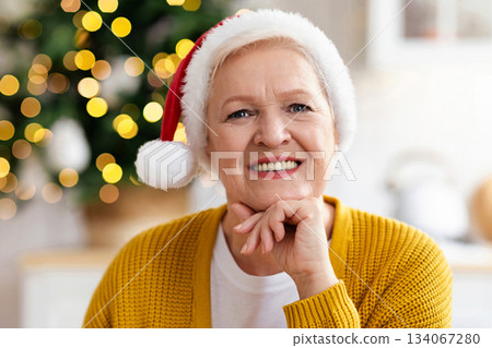 Portrait of cheerful old woman in Santa hat smiling at camera, celebrating Christmas at home, closeup. Happy senior lady sitting at kitchen decorated with xmas tree, smiling at camera Portrait of cheerful old woman in Santa hat smiling at camera, celebrating Christmas at home, closeup. Happy senior lady sitting at kitchen decorated with xmas tree, smiling at camera 134067280