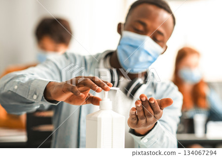 Hand Disinfection Concept. African american male student applying antibacterial sanitizer on hands from bottle, pouring it on palms, wearing protective medical mask, sitting at table at class 134067294