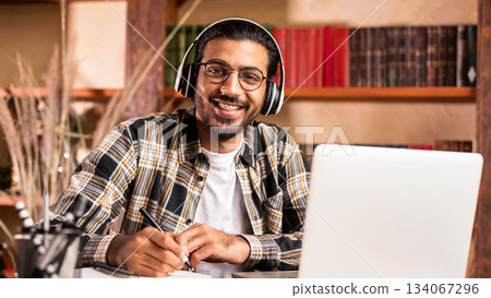 Indian Student Guy Posing At Laptop Learning Online And Smiling To Camera Sitting In Library Indoor. Distance Lecture, Remote Study, E-Learning And Modern Education Concept. 134067296