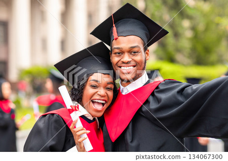Happy african american couple students in graduation dresses and hats taking selfie together, posing at university campus, enjoying and celebrating graduation, closeup portrait 134067300