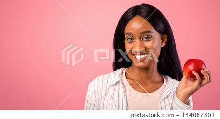 Portrait of happy black woman with tasty red apple on pink studio background, panorama. Gorgeous African American lady holding fresh delicious fruit. Healthy lifestyle and detox concept 134067301