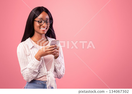 Online life. Smart black woman in glasses using cellphone for remote work or studies on pink studio background. Intelligent African American business lady having web conference on mobile device 134067302