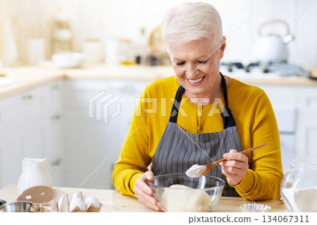 Joyful grandmother wearing apron making dough at kitchen, adding flour, copy space. Happy elderly lady baking pastry cookies or cake at home. Domestic lifestyle, culinary hobby concept 134067311