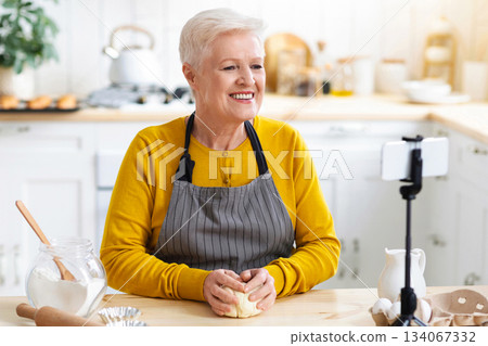 Happy senior woman chef in apron sitting at kitchen table, kneading dough and broadcasting for her followers, using mobile phone on tripod, copy space. Life while retirement concept Happy senior woman chef in apron sitting at kitchen table, kneading dough and broadcasting for her followers, using mobile phone on tripod, copy space. Life while retirement concept 134067332