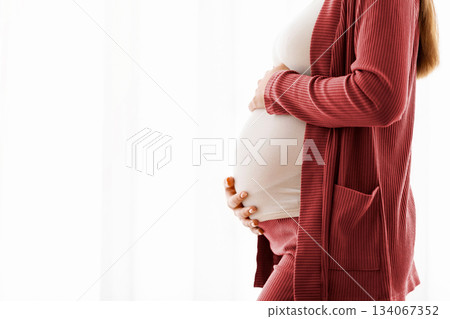 Pregnancy Concept. Happy young pregnant woman embracing belly while standing near window at home, smiling expectant mother enjoying awaiting time, side view shot with copy space, cropped Pregnancy Concept. Happy young pregnant woman embracing belly while standing near window at home, smiling expectant mother enjoying awaiting time, side view shot with copy space, cropped 134067352