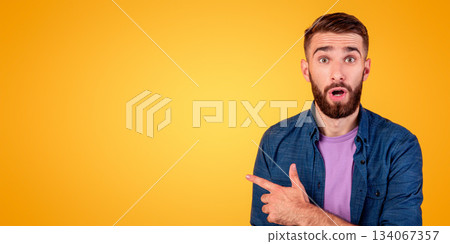Shocked millennial guy pointing aside at empty space on orange studio background, panorama. Bearded hipster guy advertising your product, offering huge sale or promo, banner design 134067357