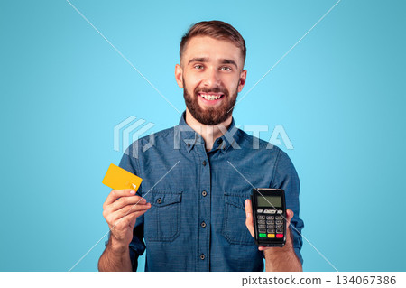 Portrait of happy young guy holding credit card and payment terminal, amiling and looking at camera over blue studio background. Positive millennial man shopping contactless during covid outbreak 134067386