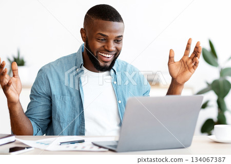Happy Black Freelancer Man Celebrating Success While Working Online On Computer, African Guy Looking At Laptop Screen, Raising Hands And Exclaiming With Excitement, Sitting At Desk In Home Office 134067387