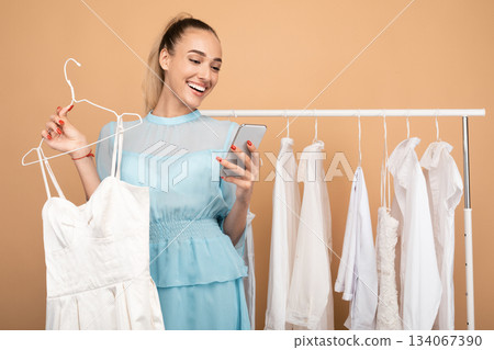 Portrait of smiling young woman using her mobile phone, holding hanger with dress, chatting and sending photos to her friend, choosing outfit, standing near clothing rail at the studio Portrait of smiling young woman using her mobile phone, holding hanger with dress, chatting and sending photos to her friend, choosing outfit, standing near clothing rail at the studio 134067390
