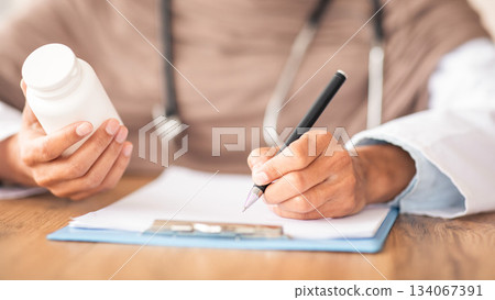 Medicine, pills, vitamins concept. Closeup of unrecognizable female doctor in hijab holding medicine, writing prescription in medical chart, prescribing treatment for patient in hospital, cropped 134067391