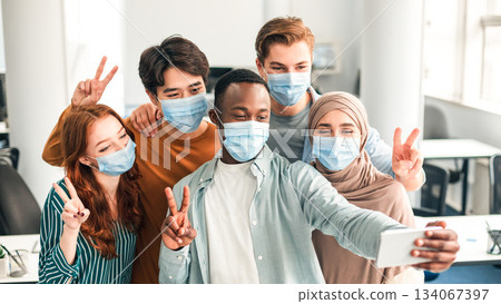 Friendship, Lifestyle And People Concept. Portrait of smiling happy diverse multiracial group wearing surgical face masks and taking selfie indoors, using mobile phone, showing peace or victory sign Friendship, Lifestyle And People Concept. Portrait of smiling happy diverse multiracial group wearing surgical face masks and taking selfie indoors, using mobile phone, showing peace or victory sign 134067397