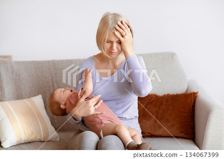 Exhausted blonde woman young mother having migraine, holding crying little baby, home interior, copy space. Tired caucasian mom having headache, touching her forehead. Stressful motherhood concept 134067399