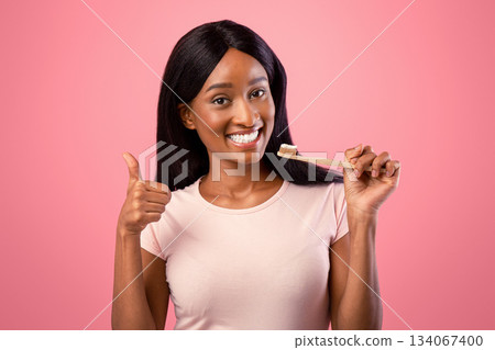 Pretty black lady with beautiful smile holding wooden toothbrush and showing thumb up gesture on pink studio background. Happy African American woman taking care of her teeth health Pretty black lady with beautiful smile holding wooden toothbrush and showing thumb up gesture on pink studio background. Happy African American woman taking care of her teeth health 134067400
