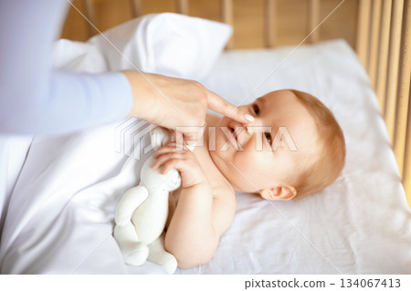 Smiling little kid laying in wooden baby bed under blanket, holding toy, mother finger touching happy kid nose, unrecognizable loving mom playing with awaken cute toddler, closeup 134067413