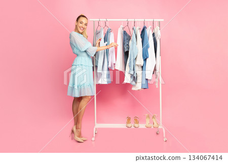 Full length vertical portrait of cheerful smiling woman standing at boutique wardrobe, choosing fashion stylish clothes at rack with hangers, looking at camera, looking for outfit, pink background 134067414