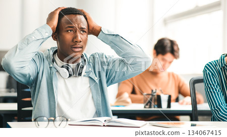 Portrait of shocked black student sitting at desk in classroom, grabbing his head. Sad African American youth unprepared for test or exam, thinking about deadline or hard new theme Portrait of shocked black student sitting at desk in classroom, grabbing his head. Sad African American youth unprepared for test or exam, thinking about deadline or hard new theme 134067415