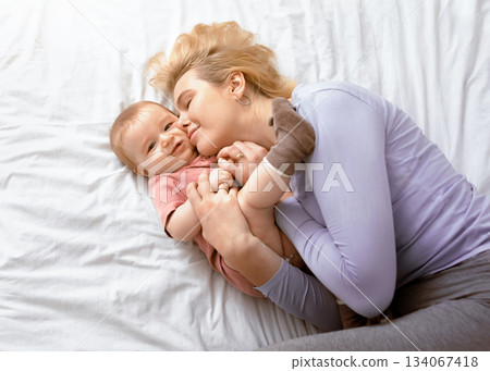 Love and tenderness, motherhood concept. Loving blonde mother caressing her cute baby boy, lying together on bed, top view. Young caucasian woman bonding with little kid at home, copy space 134067418
