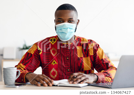 Portrait Of African Man In Medical Mask And Traditional Clothes Sitting At Desk In Home Office, Black Freelancer Guy Working Remotely During Coronavirus Pandemic, Looking At Camera, Copy Space Portrait Of African Man In Medical Mask And Traditional Clothes Sitting At Desk In Home Office, Black Freelancer Guy Working Remotely During Coronavirus Pandemic, Looking At Camera, Copy Space 134067427