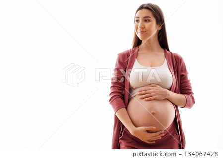 Young beautiful pregnant woman standing near window at home and touching belly, happy future mother tenderly embracing her naked tummy and looking away, enjoying pregnancy time, copy space 134067428