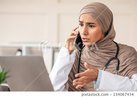 Emotional female doctor young lady in hijab looking at laptop screen, talking on phone and gesturing, having conversation with patient, giving recommendations, working at clinic 134067434