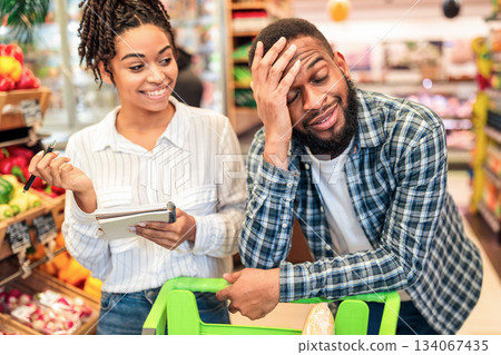 African Husband Waiting Wife Tired Of Grocery Shopping Buying Food Together Standing With Cart Full Of Products In Modern Supermarket Store. Family Of Customers Buying Groceries African Husband Waiting Wife Tired Of Grocery Shopping Buying Food Together Standing With Cart Full Of Products In Modern Supermarket Store. Family Of Customers Buying Groceries 134067435