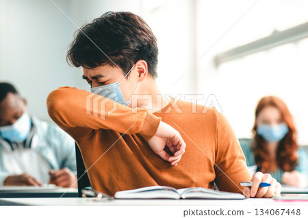 Stop Spreading Virus Concept. Asian male student couching in his elbow, sneezing in sleeve during lecture in university. Guy wearing protective medical mask, sitting at desk in classrom Stop Spreading Virus Concept. Asian male student couching in his elbow, sneezing in sleeve during lecture in university. Guy wearing protective medical mask, sitting at desk in classrom 134067448