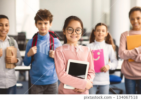 Pupils, Technology And Learning Concept. Smiling asian girl in eyeglasses holding digital tablet and standing with group of multicultural classmates in the background, studying at elementary school 134067518