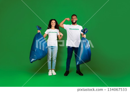 Two Diverse Volunteers Holding Blue Garbage Bags Showing Biceps And Pointing At Volunteer Inscription On T-Shirts Posing Over Green Studio Background. Keep Environment Clean. Full Length 134067533